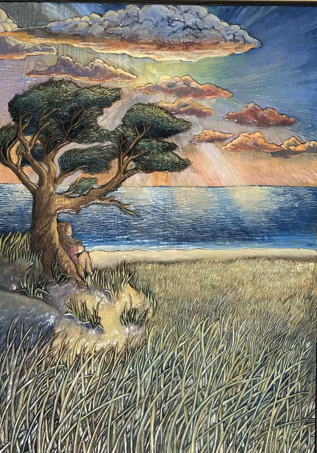 Dune Time by Gary Cota is a painted bas-relief wood carving featuring a windswept tree overlooking a Cape Cod beach. A solitary figure sits beneath the tree as sunrise illuminates colorful clouds above the ocean and marsh grasses in the foreground.